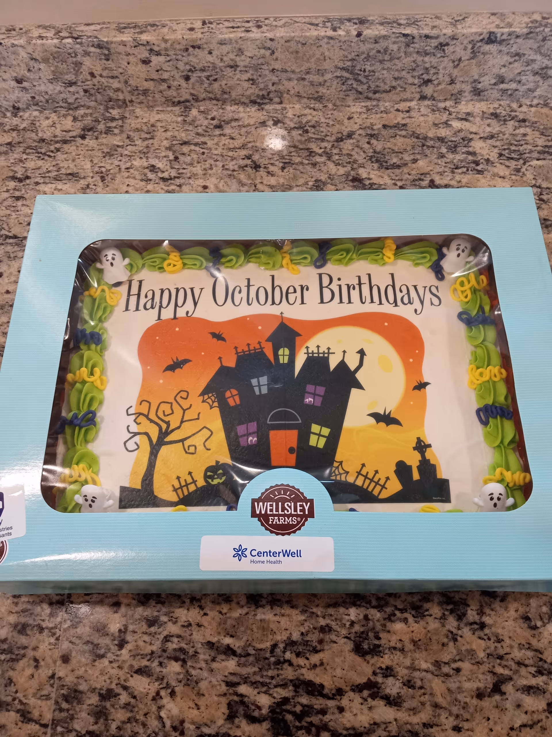 A Halloween-themed birthday cake in a light blue box on a granite countertop. The cake has green, yellow, and purple icing decorations with small ghost figures on the corners. The cake's surface features a haunted house, bats, a full moon, a tree, and a pumpkin with the text 'Happy October Birthdays'.