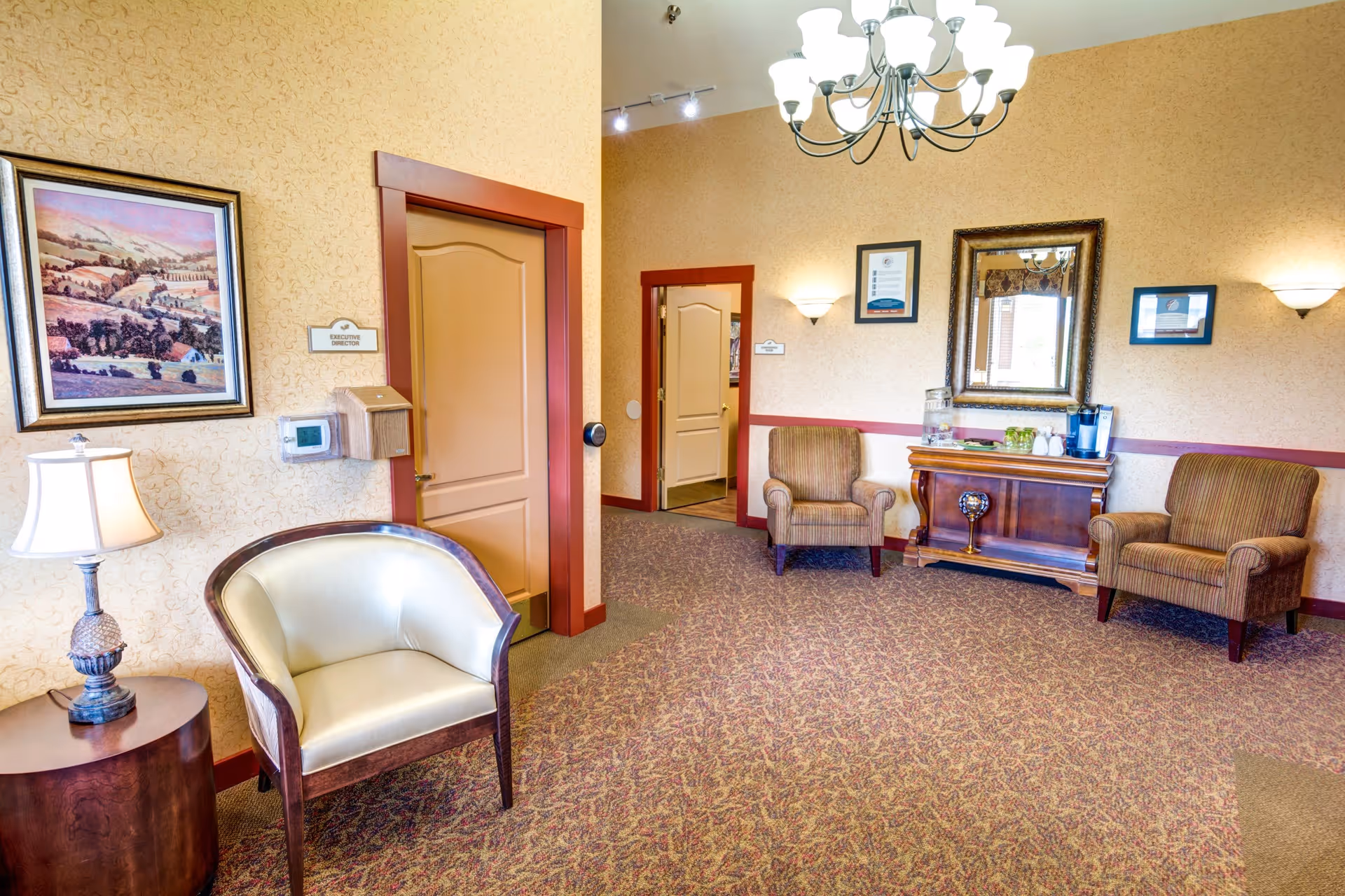 A cozy waiting area with three armchairs, a wooden side table with a lamp, a wooden console table with a water dispenser and cups, a large mirror, framed pictures on the walls, and a chandelier hanging from the ceiling. The walls are beige with a red trim, and there are two closed doors visible.