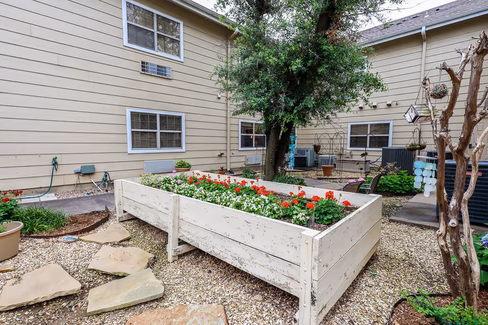 Outdoor garden area at Aberdeen Heights Assisted Living featuring a raised white wooden planter box filled with red and white flowers. The garden is surrounded by gravel and stone stepping paths, with a tree and several potted plants nearby. The beige exterior walls of the building with windows and air conditioning units are visible in the background.