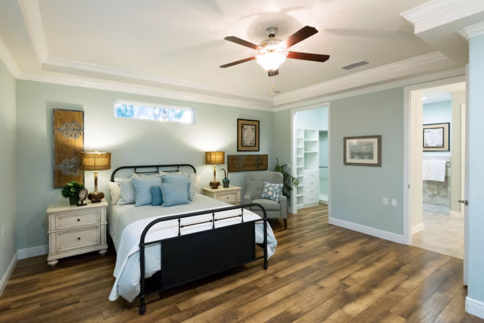 A bright and spacious bedroom with light blue walls, a ceiling fan with light, and wood flooring. The room features a black metal bed frame with white and blue bedding and multiple pillows. There are two white nightstands with matching lamps on either side of the bed. A gray upholstered chair with a patterned pillow is placed near an open doorway leading to a walk-in closet and a bathroom. The bathroom has beige tiles and a towel hanging on the wall. The room is decorated with framed artwork and decorative wall panels.