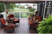 Covered porch with wooden floor featuring four cushioned wicker chairs arranged around a small table, potted plants on the railing and floor, overlooking a green lawn and trees in the background.