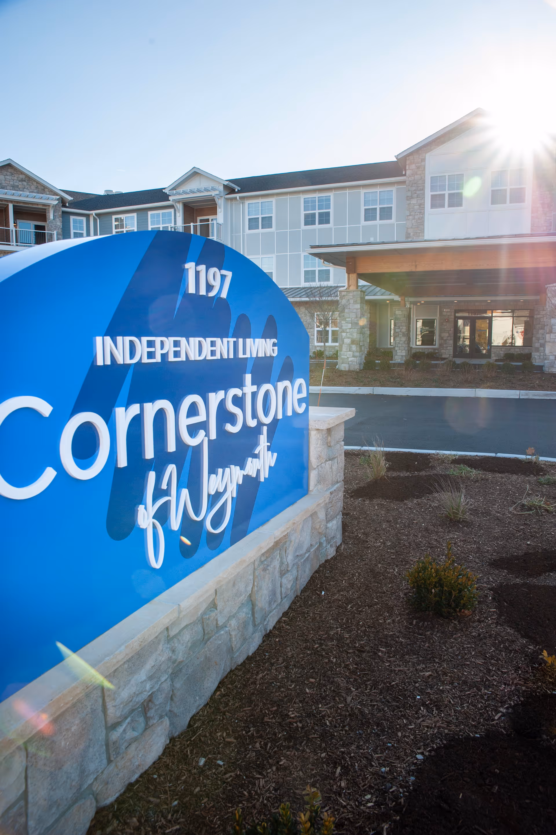 Blue entrance sign reading 'Cornerstone of Weymouth Independent Living' in front of the facility building with sunlight.