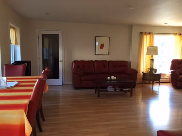 Spacious living room with red leather sofas, a coffee table, a dining table with red chairs and striped tablecloth, hardwood floors, and curtained windows.