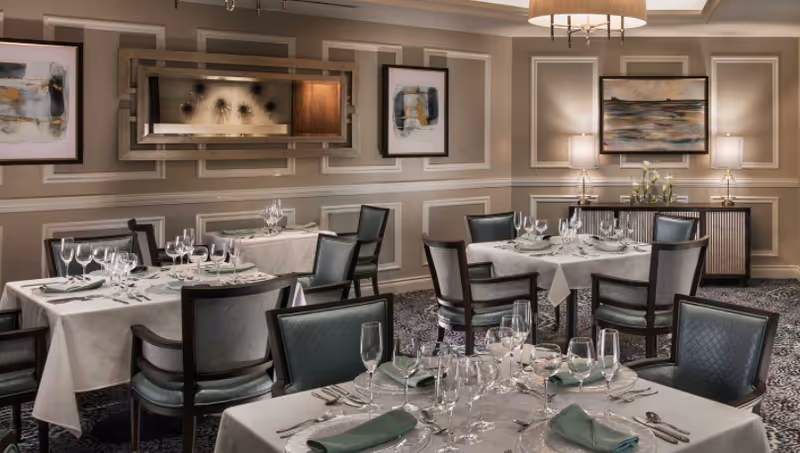 Elegant dining room with multiple tables covered in white tablecloths, set with glassware, silverware, and green napkins. The room features patterned carpet, paneled walls with framed artwork, a large mirror, and soft lighting from table lamps and a ceiling fixture.
