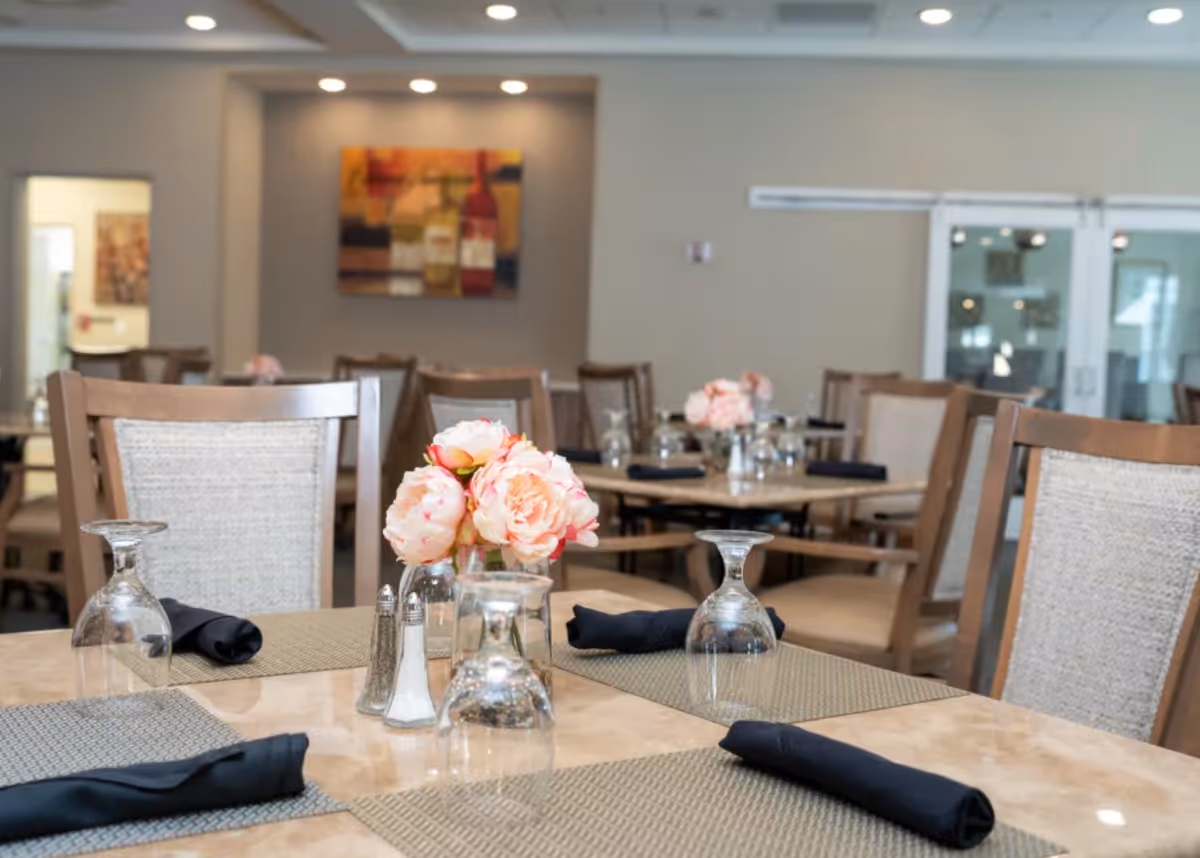 Dining area with wooden chairs and tables set with placemats, napkins, upside-down wine glasses, and small floral centerpieces.