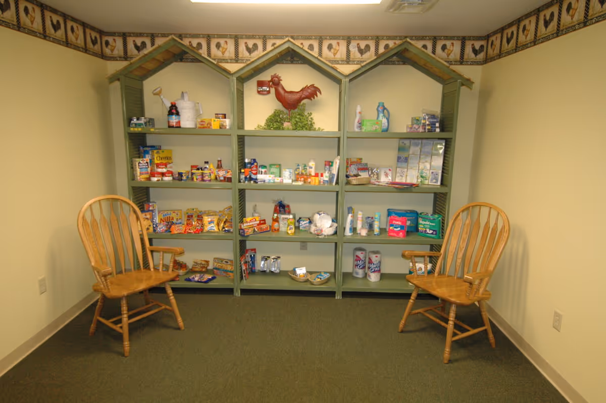 A small interior room with house-shaped green shelving stocked with snacks and household items, flanked by two wooden chairs.