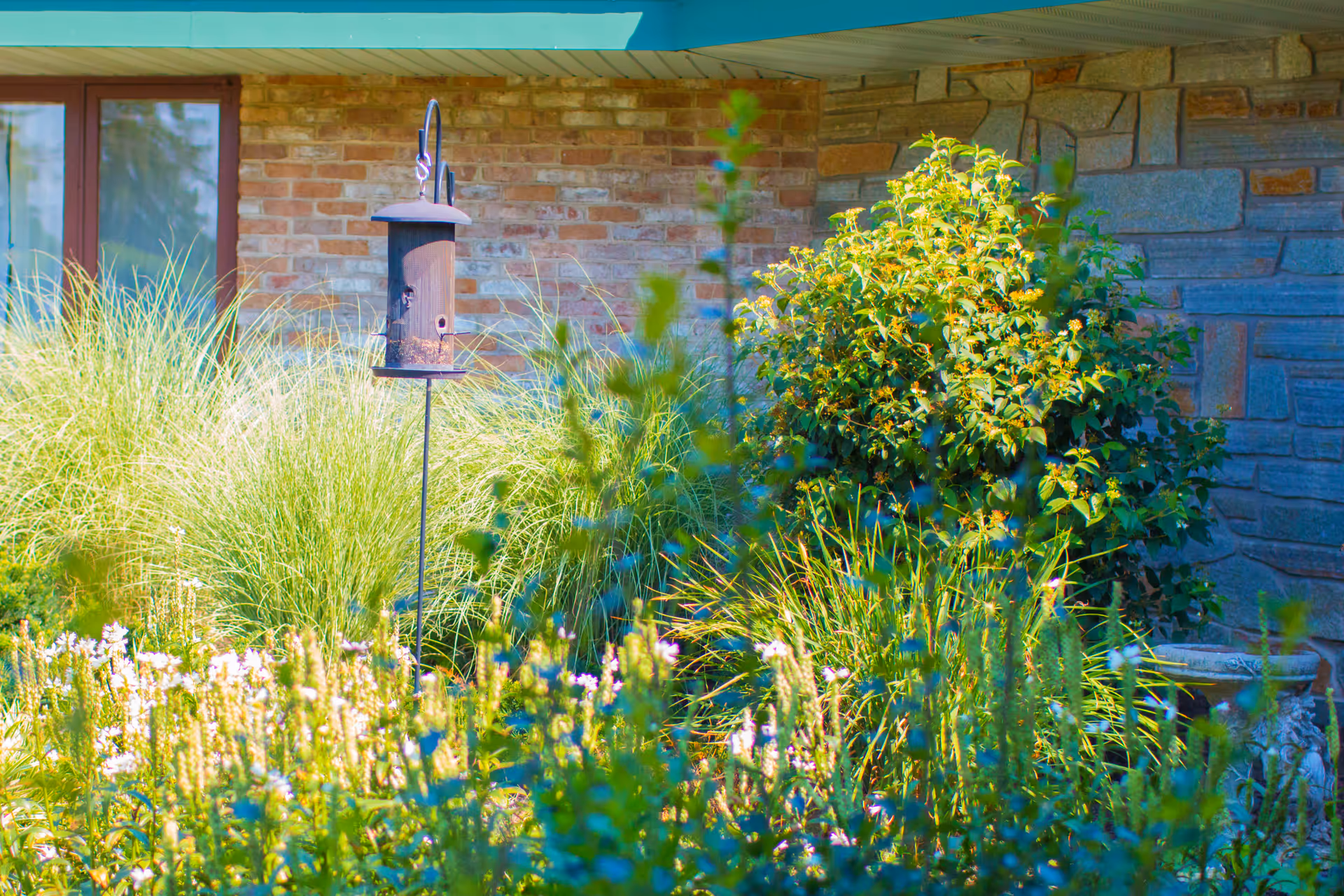 A garden area with various green plants and flowers in front of a brick and stone building wall. A bird feeder is mounted on a pole among the plants.