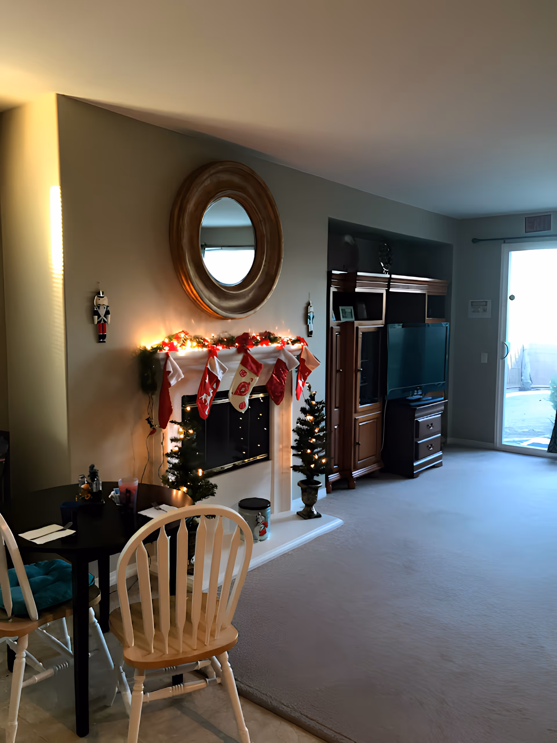 A cozy living room decorated for Christmas with a fireplace adorned with stockings and garlands with lights. Two small Christmas trees with lights flank the fireplace. Above the fireplace is a large round mirror. To the right, there is a wooden entertainment center with a TV and a sliding glass door leading outside. In the foreground, there is a small dining table with two wooden chairs.