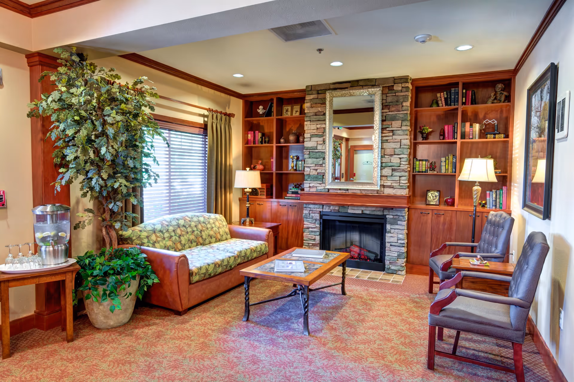 A cozy living room area in Washington Gardens Memory Care featuring a patterned sofa, two leather armchairs, a wooden coffee table, a stone fireplace with a large mirror above it, built-in wooden bookshelves filled with books and decorative items, a large potted plant, a water dispenser on a small table, and warm lighting from table lamps.