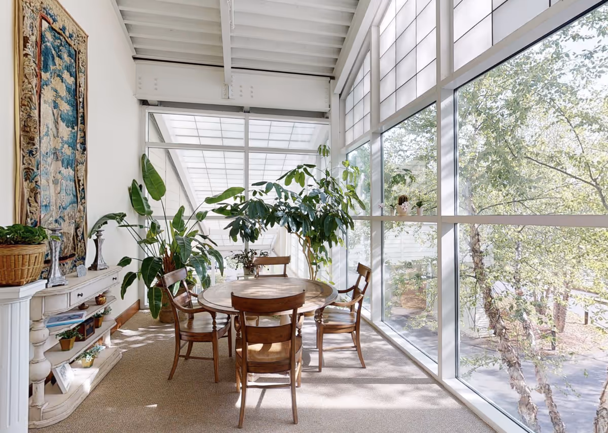 A bright sunroom with large floor-to-ceiling windows showing trees outside. Inside, there is a round wooden table surrounded by four wooden chairs. Several green potted plants are placed around the room, and a white decorative console table with small plants and decorative items is against the wall. A tapestry hangs on the wall above the console table.