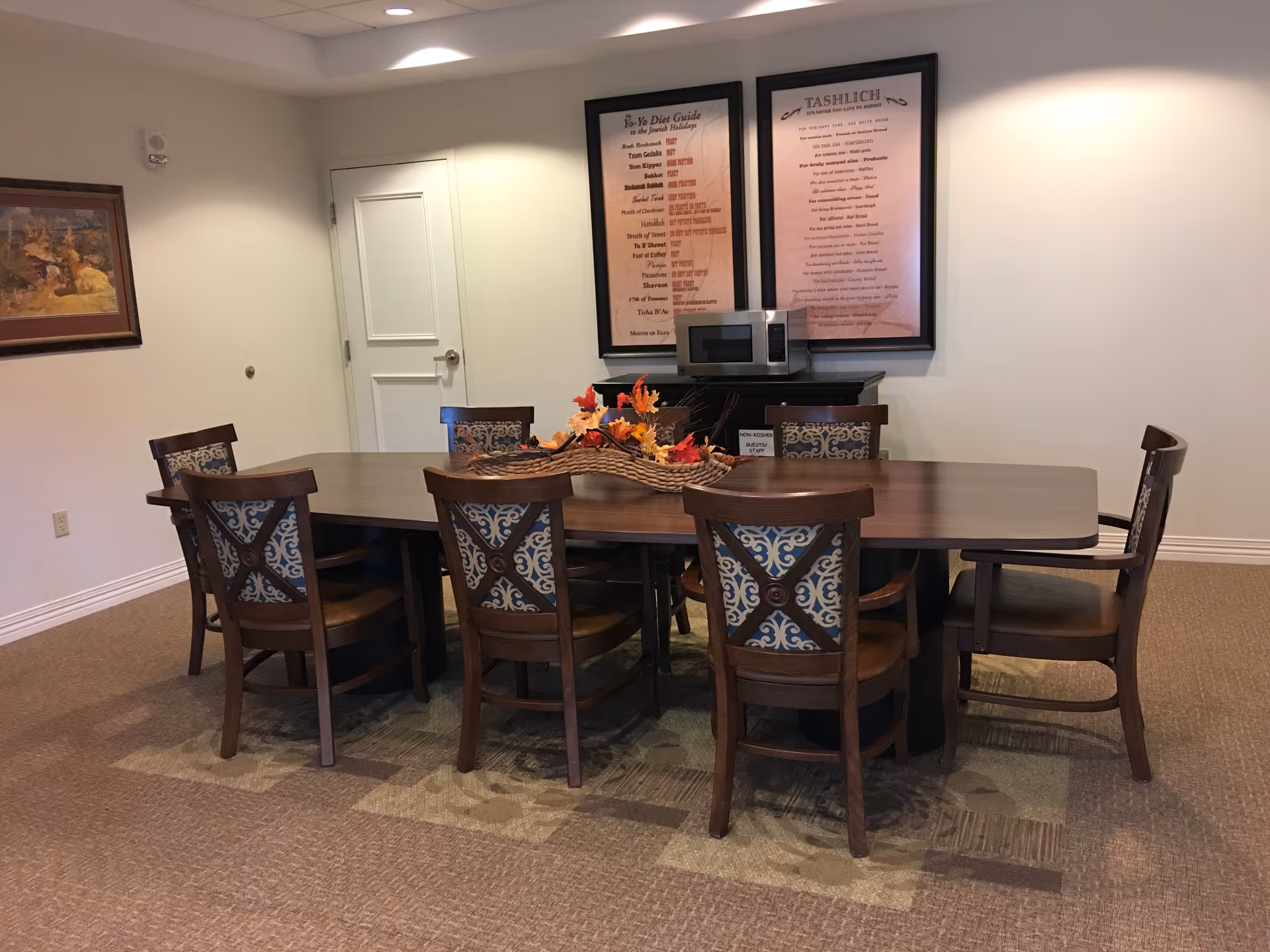 A communal dining room with a long wooden table surrounded by patterned chairs and a fall centerpiece, with framed artwork and a microwave on a sideboard against the wall.