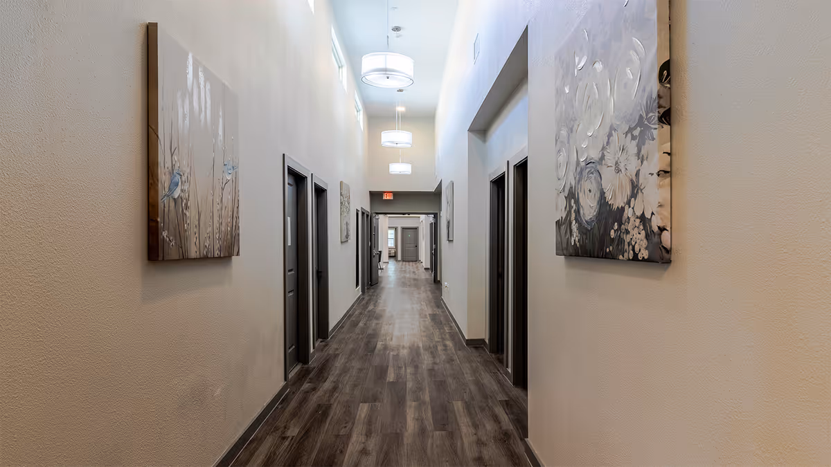 A long, well-lit hallway in a senior living facility with wooden flooring and beige walls. Several closed doors line both sides of the hallway, and decorative paintings hang on the walls. Modern ceiling lights hang from the high ceiling, and an exit sign is visible at the far end.