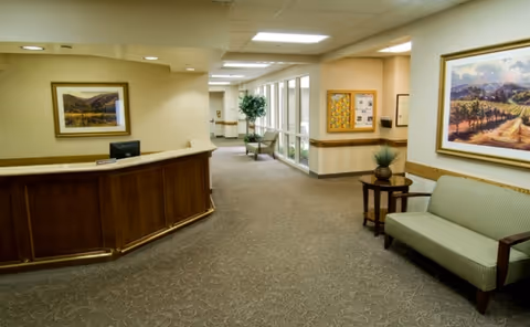 Interior view of a senior living facility hallway with a wooden reception desk on the left, a green cushioned bench with wooden arms on the right, framed landscape paintings on the walls, a small table with a plant, and a windowed wall with chairs and a potted plant further down the corridor.