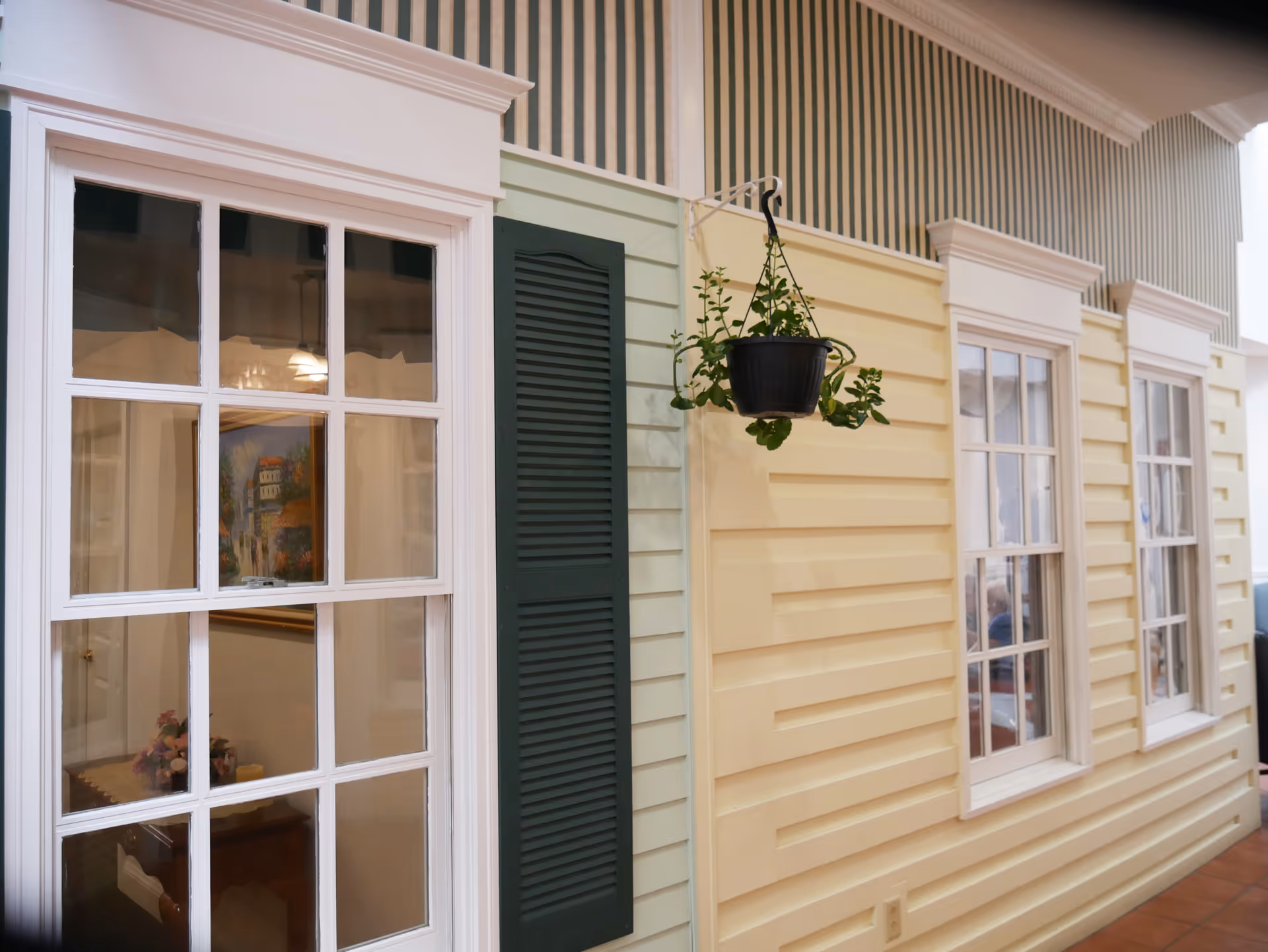Interior hallway with a faux house facade showing cream siding, white-framed windows, a green shutter and a hanging potted plant.