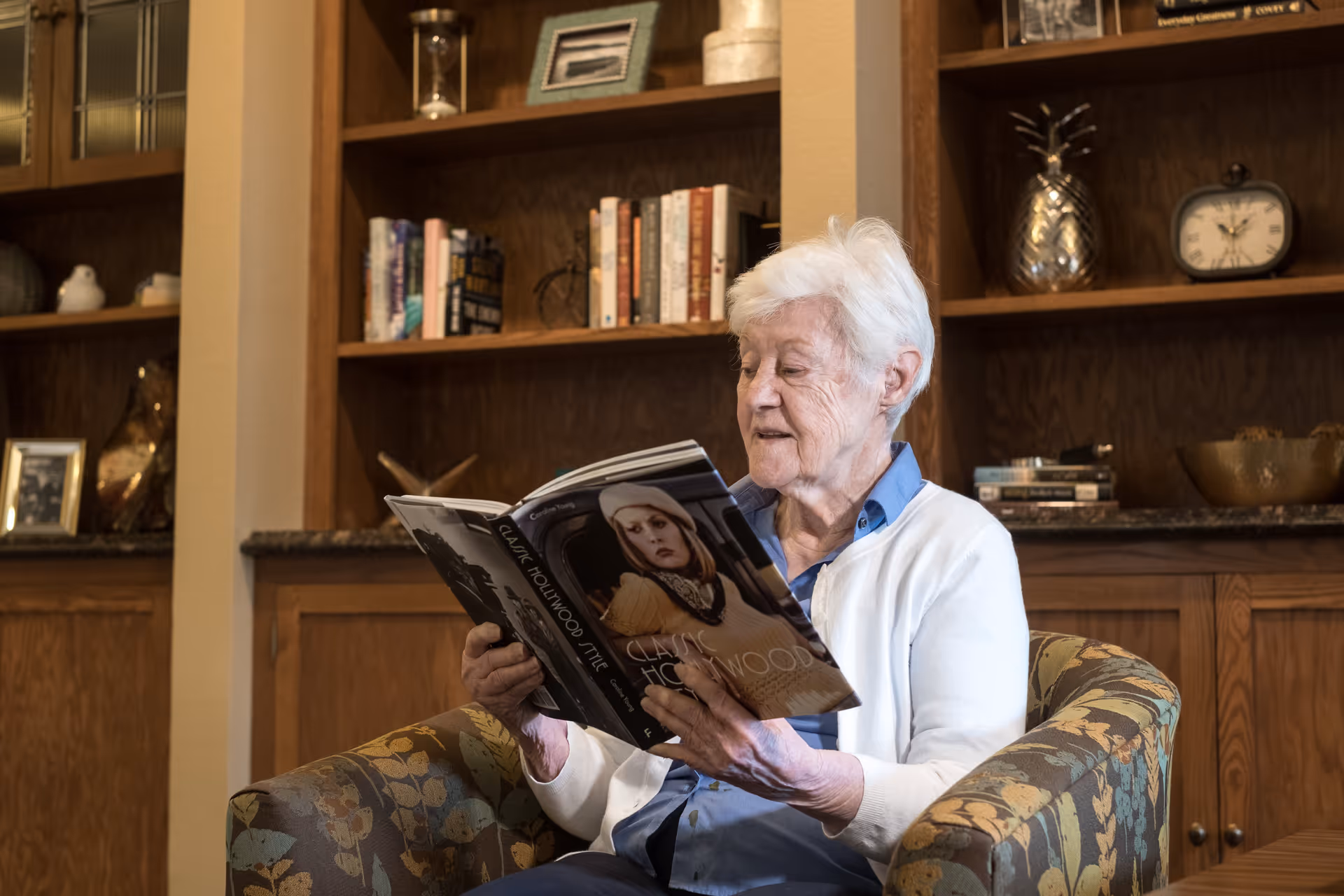 An elderly woman with white hair sitting in a floral patterned armchair, reading a book titled 'Classic Hollywood'. Behind her is a wooden bookshelf filled with books and decorative items including a clock and a silver pineapple.