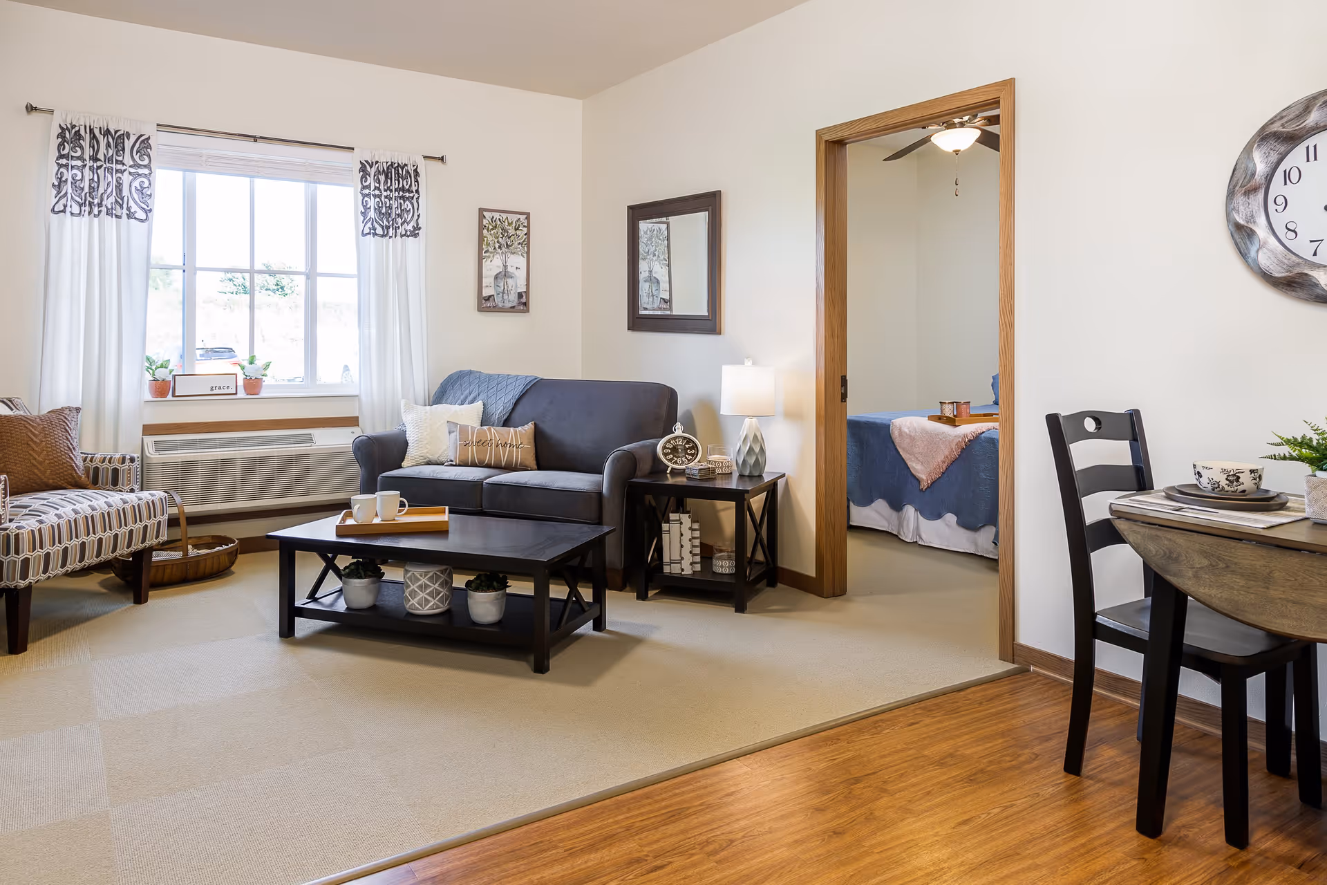 A cozy living room with a dark blue loveseat, patterned armchair, and a black coffee table with decorative plants underneath. A window with white curtains and patterned valances lets in natural light. A side table with a lamp and clock sits next to the loveseat. A doorway leads to a bedroom with a bed covered in blue and pink bedding. A small dining table with two chairs is partially visible on the right.
