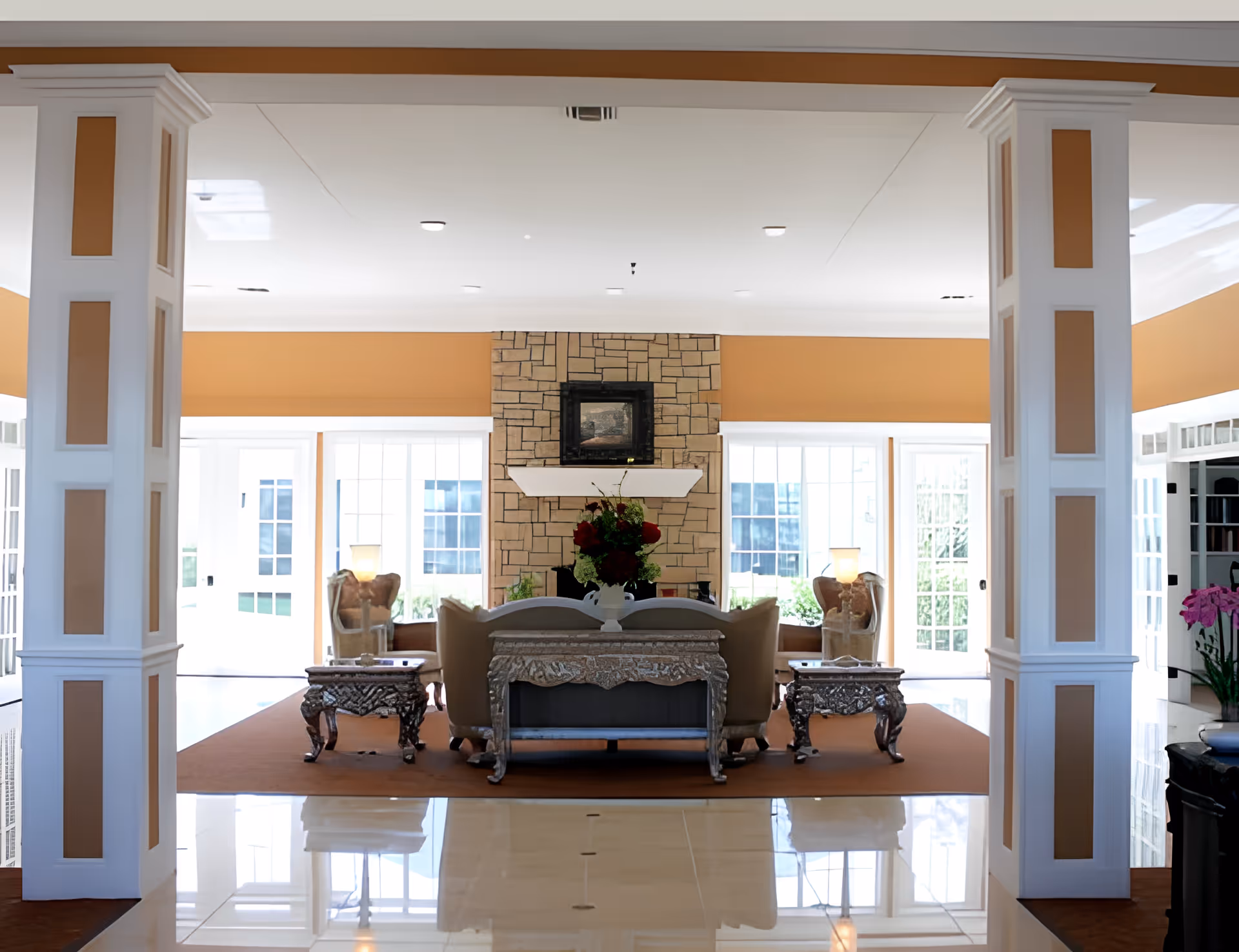 A bright and spacious living room area in Silver Creek Assisted Living featuring a stone fireplace with a painting above it, surrounded by large windows letting in natural light. The room includes elegant furniture such as a sofa, armchairs, two ornate side tables, and a decorative console table with a floral arrangement. The walls are painted in warm tones, and the floor is shiny and reflective.