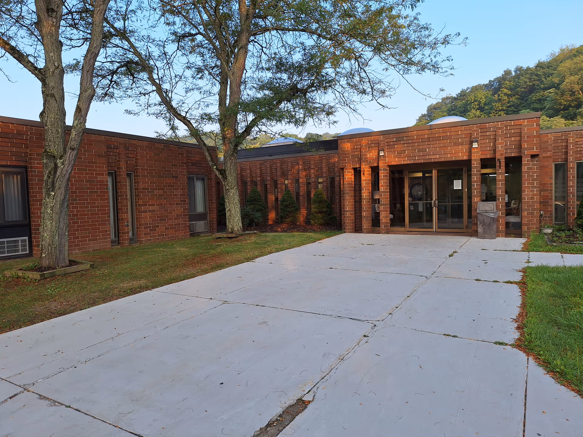 Exterior view of a single-story brick building with large windows and a glass entrance door. There are trees and shrubs near the building, and a concrete pathway leads to the entrance. The sky is clear and blue.