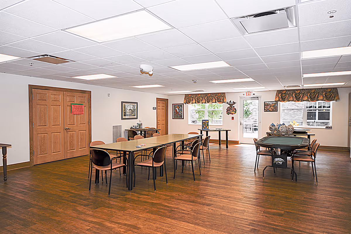 A spacious room with wooden flooring and white ceiling tiles, featuring two rectangular tables surrounded by chairs. The room has large windows with floral valances, a door with an exit sign above it, and framed pictures on the walls. There are some decorative items on one of the tables and a double wooden door on the left side of the room.