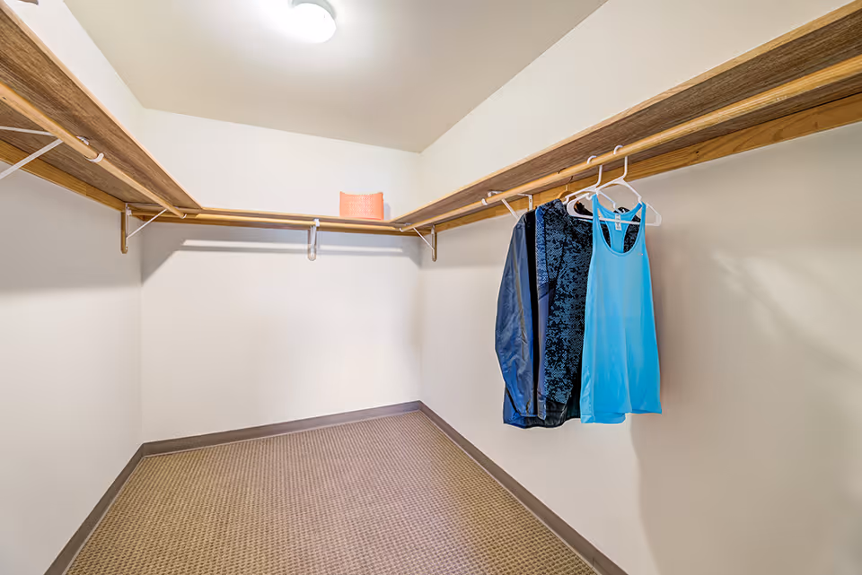 A spacious walk-in closet with beige carpet flooring, white walls, and wooden shelves with hanging rods. There are three pieces of clothing hanging on the right side and a folded orange towel placed on the back shelf under a ceiling light.