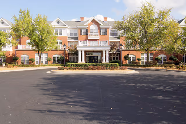 Front exterior view of a large, three-story brick building with white columns at the entrance, surrounded by trees and landscaping under a partly cloudy sky.