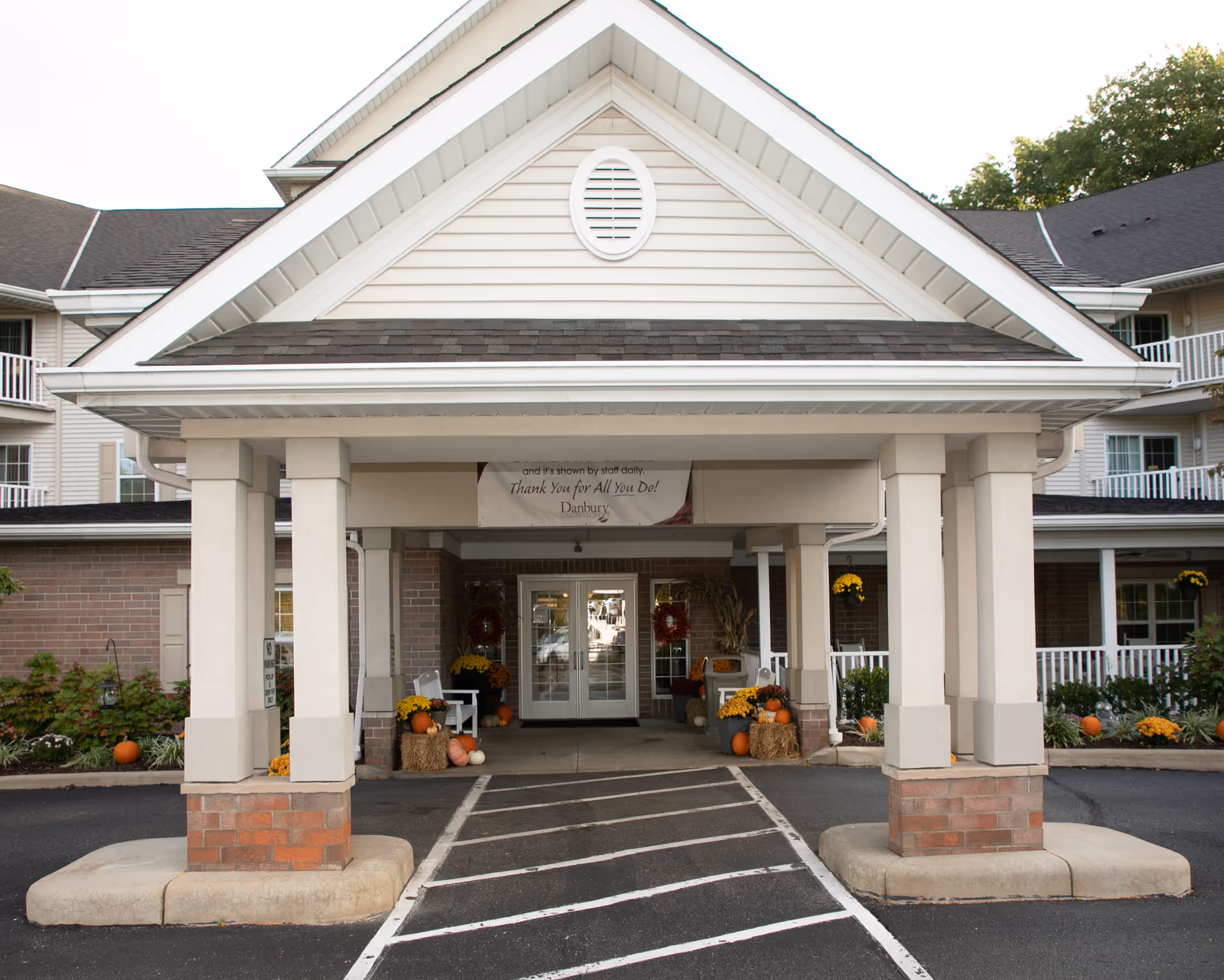 Entrance of a senior living facility with a covered driveway supported by columns. The entrance is decorated with pumpkins, hay bales, and yellow flowers, suggesting a fall or autumn theme. The building has beige siding and brick accents with white railings on the balconies.