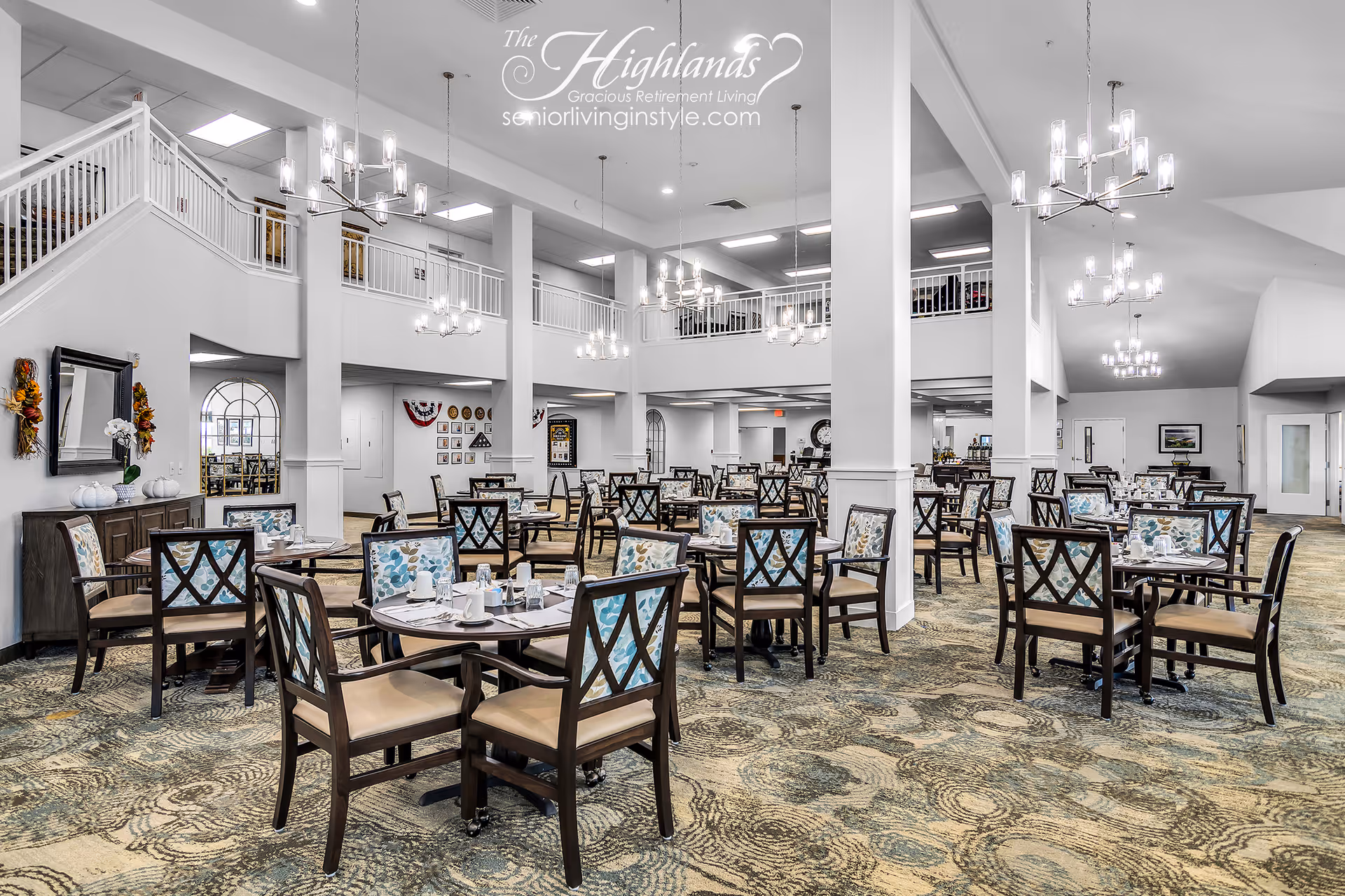 Spacious and well-lit dining room in a retirement living facility with multiple round tables and chairs arranged neatly. The chairs have patterned backs and cushioned seats. The room features high ceilings with modern chandeliers, a carpeted floor with a swirling pattern, and a second-floor balcony overlooking the dining area. Walls are decorated with framed pictures and seasonal wreaths.