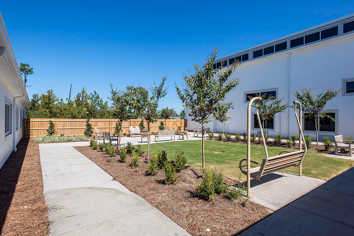 Sunny courtyard with paved paths, lawn, benches, young trees, and a wooden fence beside a white building.