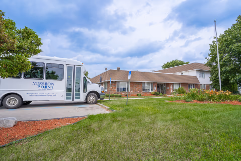 A Mission Point shuttle bus parked outside the front of a single-story brick nursing and rehabilitation building under a cloudy sky.