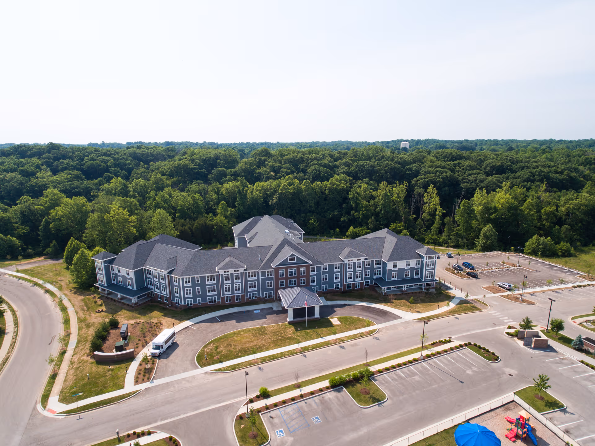 Aerial view of Evergreen Village at Bloomington, showing a large, modern senior living facility surrounded by trees and greenery. The building has a gray and white exterior with multiple windows and a covered entrance. There are parking lots with several cars and a playground with a blue umbrella visible in the lower right corner.