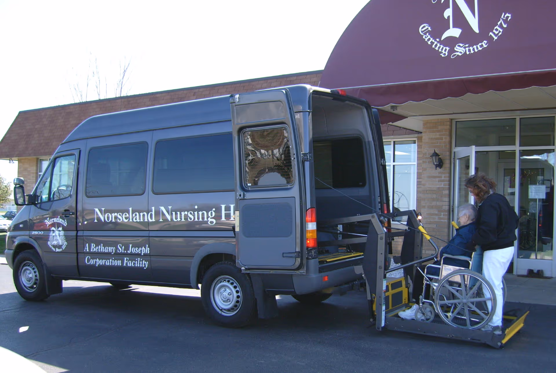 A dark-colored Norseland Nursing Home van with its rear door open and a wheelchair lift deployed. A person in a wheelchair is being assisted by a caregiver onto the lift outside the entrance of a building with a maroon awning.