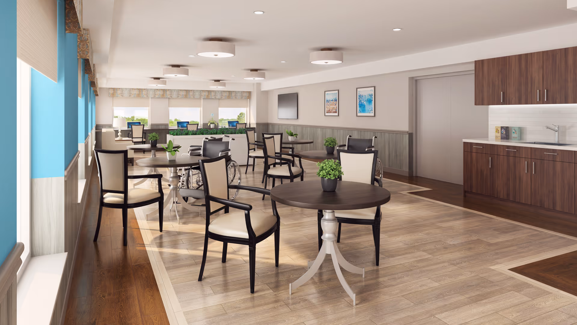 Bright communal dining area with round tables, chairs, potted plants, a kitchenette and large windows.