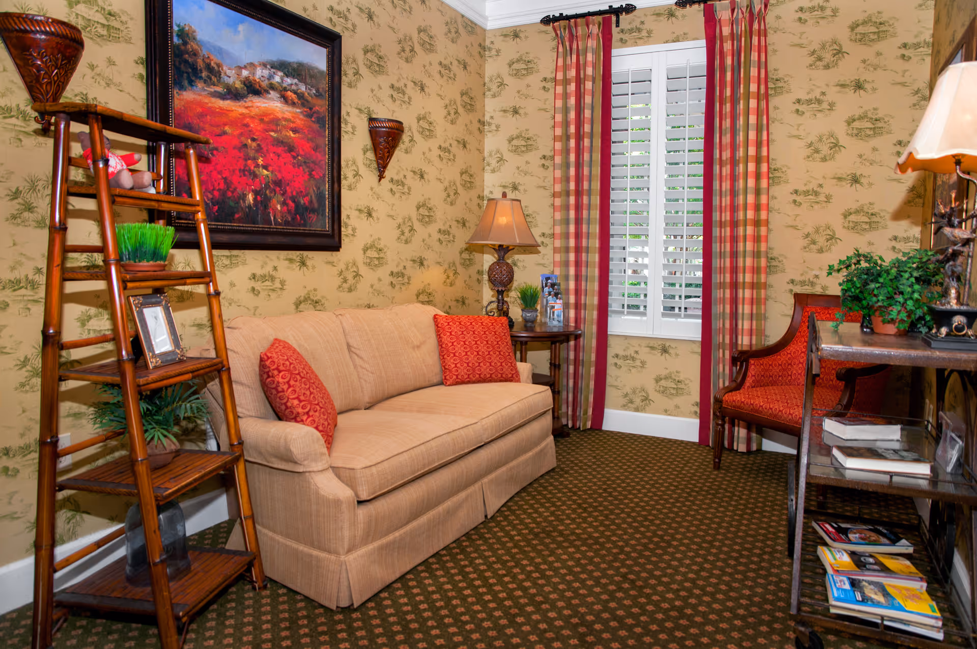 A cozy living room with a beige sofa adorned with two red patterned cushions, a wooden ladder-style shelf holding decorative items, a side table with a lamp and small plants, a red upholstered chair, and a window with white shutters and red plaid curtains. The walls have a yellowish wallpaper with green patterns, and a framed painting of a red flower field hangs above the sofa.