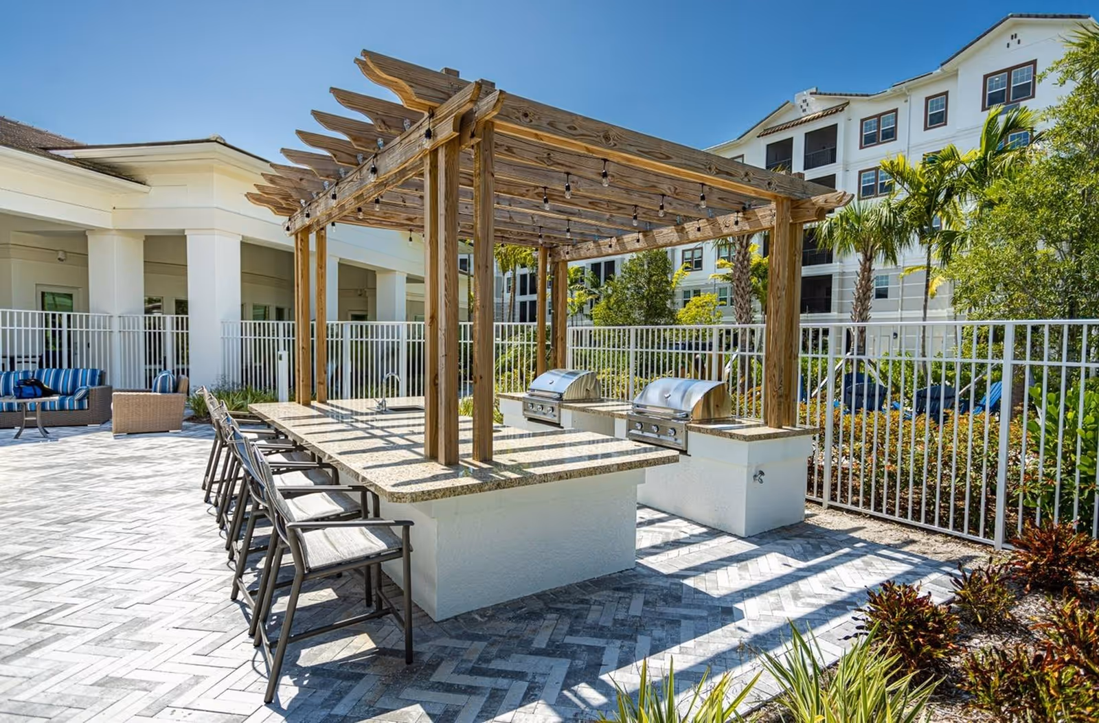 Outdoor communal grilling area with a wooden pergola, bar seating and two stainless-steel grills in front of an apartment building.