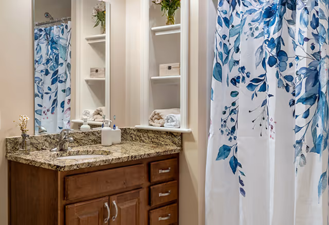 Bathroom with a granite countertop vanity featuring a sink and faucet, wooden cabinets with silver handles, a large mirror above the sink, open shelves with towels and decorative items, and a shower with a white curtain decorated with blue and green leafy patterns.