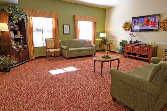 A cozy living room with green sofas, a wooden coffee table with a sunflower decoration, a wooden TV stand with a flat-screen TV mounted on the wall, two windows with orange curtains, a wooden cabinet with shelves, and various plants and lamps providing warm lighting.
