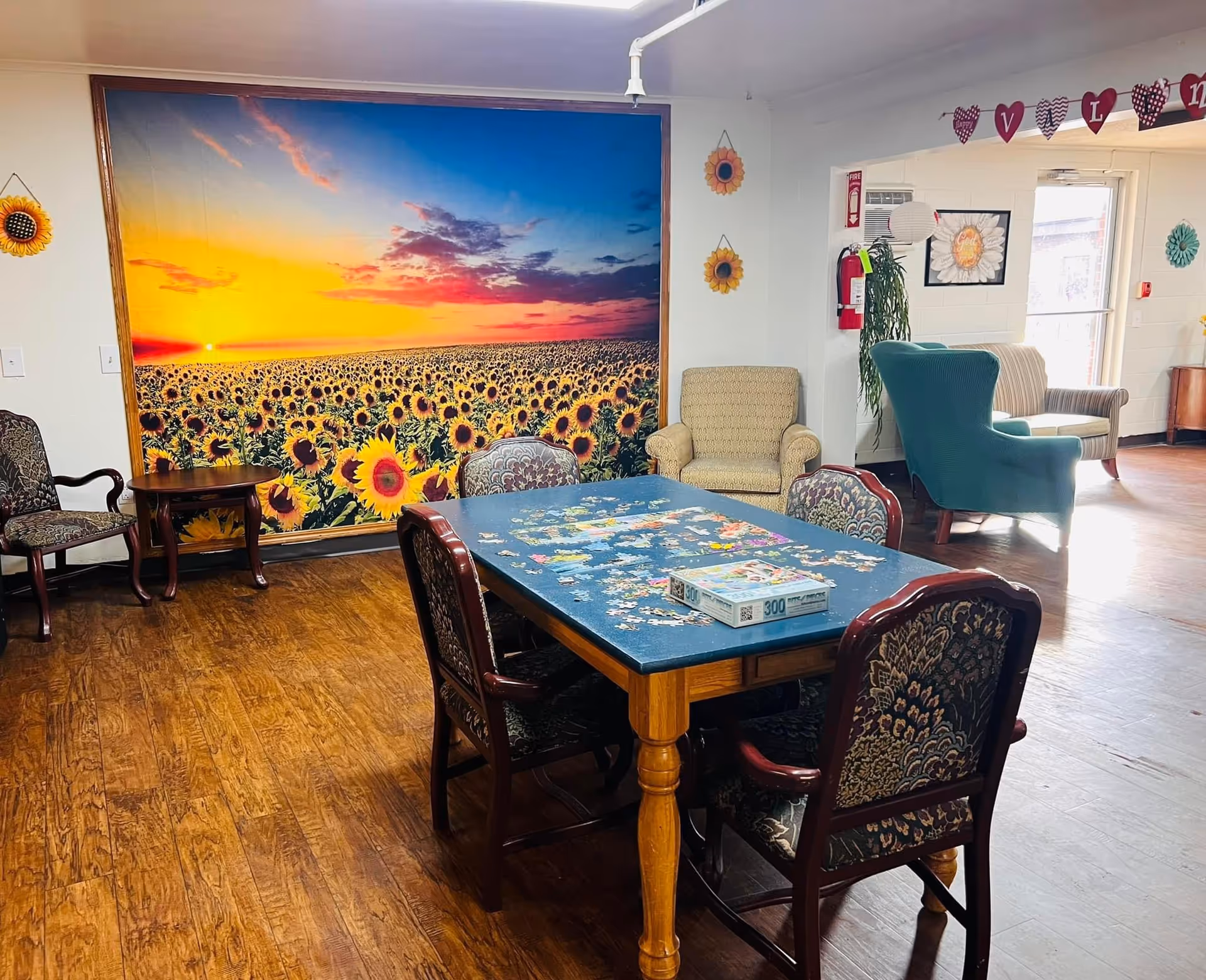 A cozy communal activity room with a puzzle-covered table surrounded by upholstered chairs, lounge seating, and a large sunflower-sunset mural on the wall.