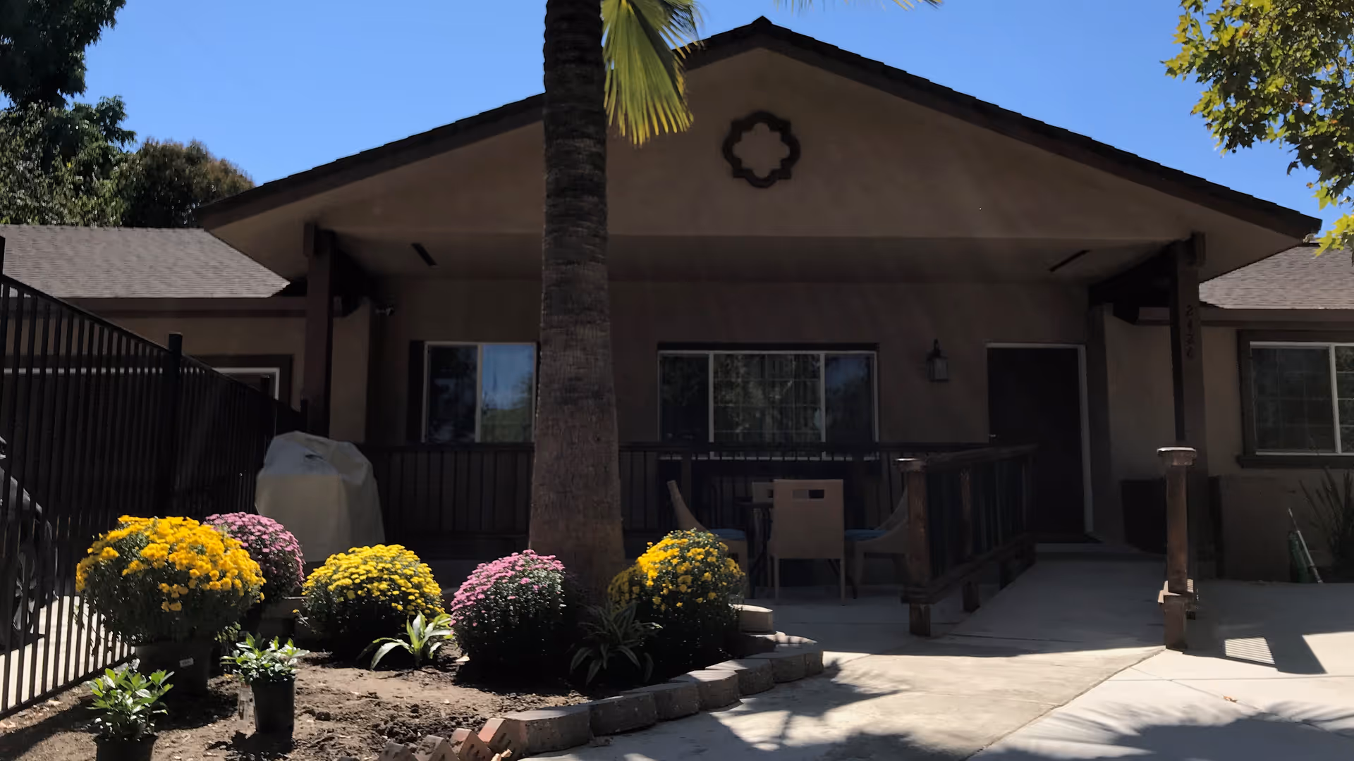Front exterior view of a single-story residential building with a covered porch, a palm tree in the center, and colorful flower bushes planted in front. There is a black metal fence on the left side and outdoor furniture on the porch.