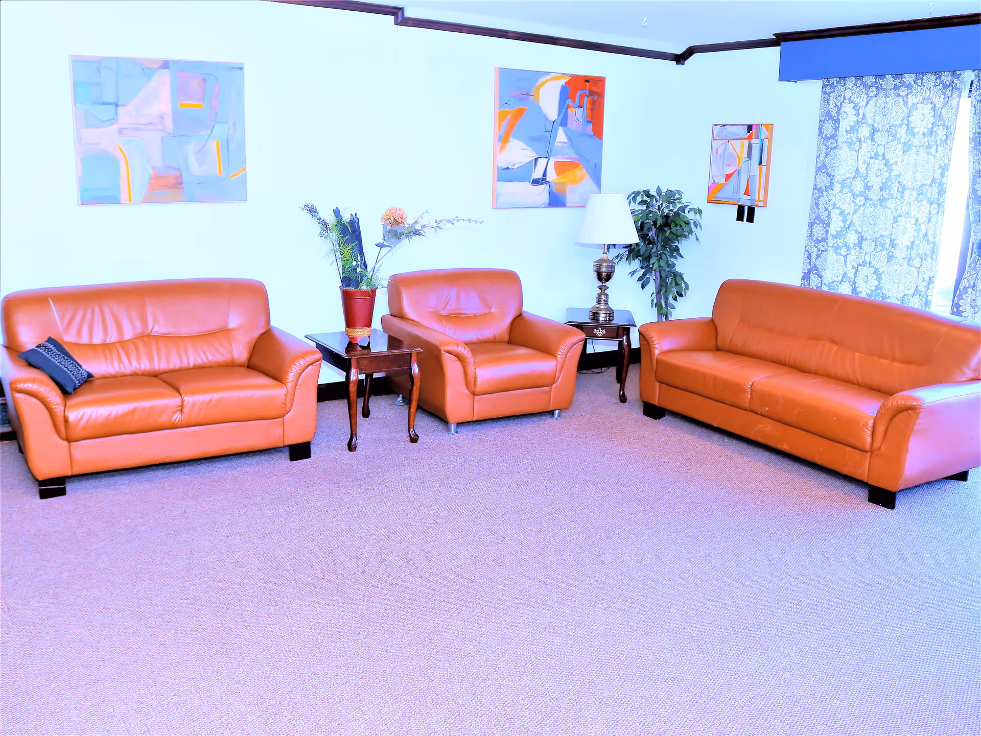 A living room with two orange leather sofas and one matching armchair arranged around two wooden side tables. One side table has a flower vase with artificial flowers, and the other has a lamp. The walls are decorated with three abstract paintings, and there is a window with patterned curtains on the right side.