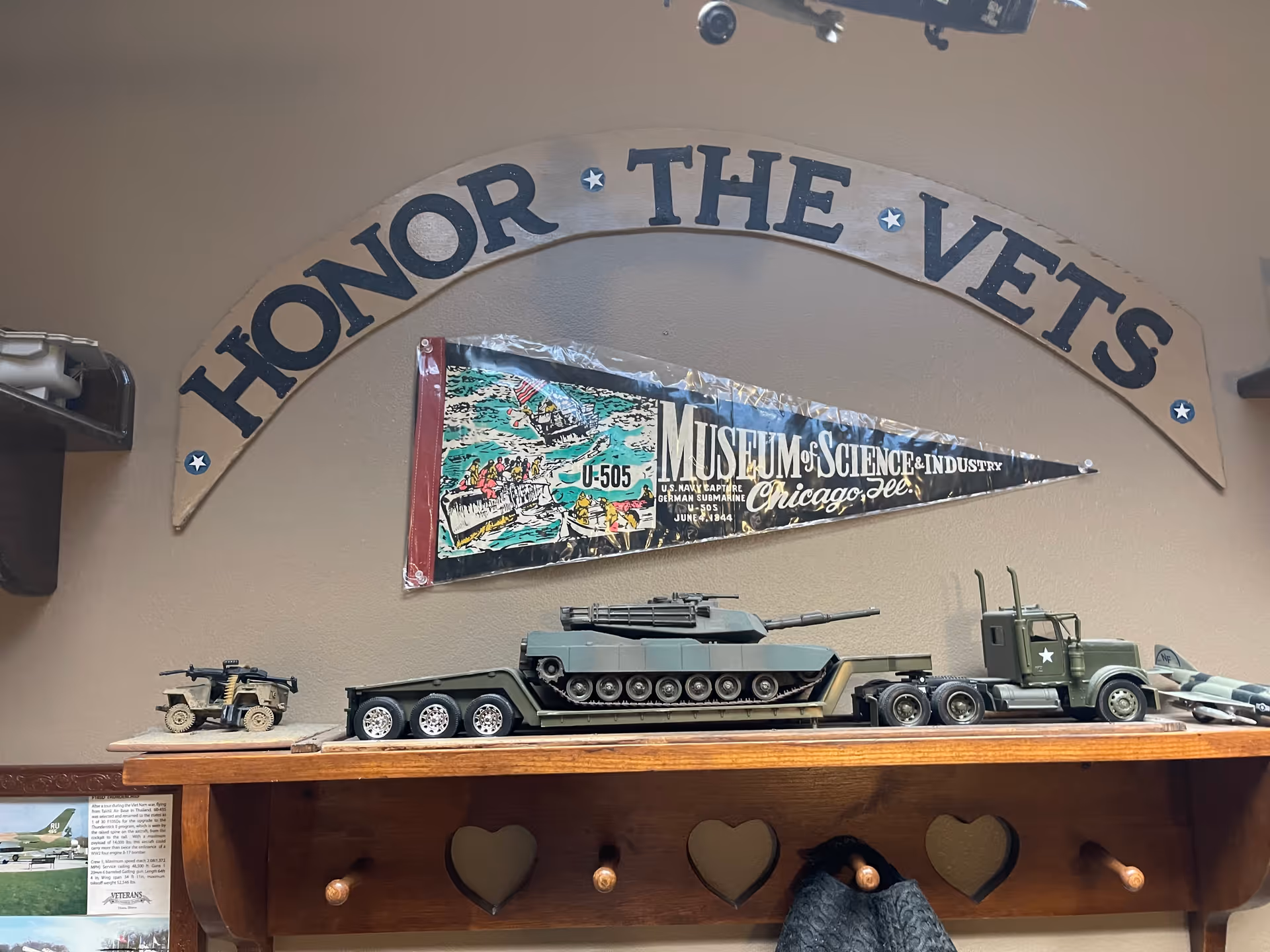 A wall display featuring a wooden arch with the words 'HONOR THE VETS' above a pennant from the Museum of Science & Industry in Chicago, Illinois. Below the pennant is a model military tank on a flatbed trailer attached to a green military truck, with a small military jeep model to the left. The display is mounted on a wooden shelf with heart-shaped cutouts and pegs, one of which holds a dark-colored hat.