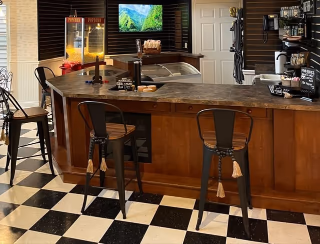 Interior view of a snack bar area with a checkered black and white floor. There is a wooden counter with three metal bar stools that have wooden seats and tassels hanging from the backrests. Behind the counter, there is a popcorn machine, a television mounted on a dark wooden wall, and various shelves holding coffee supplies and condiments. A small sign with pub rules is visible on the right side of the counter.