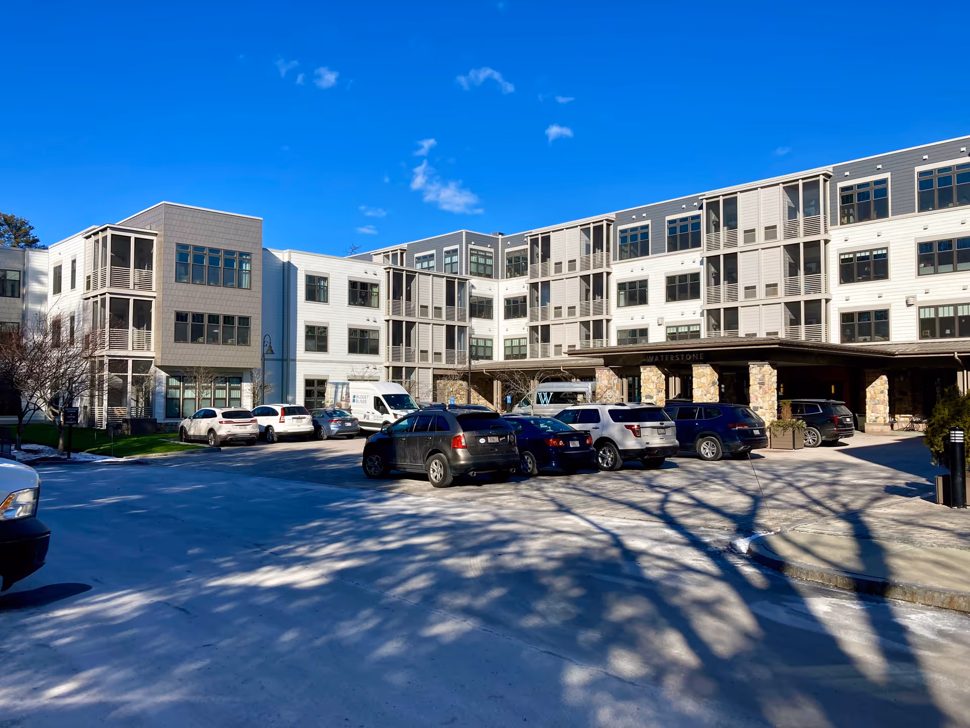 Exterior view of Waterstone At Wellesley, a multi-story senior living facility with a parking lot in front. Several cars are parked, and the building has large windows and a covered entrance supported by stone pillars. The sky is clear and blue.