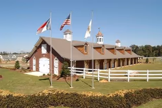 Long single-story brick and wood building with cupolas, multiple flagpoles, and a white fence on a manicured lawn under a clear sky.