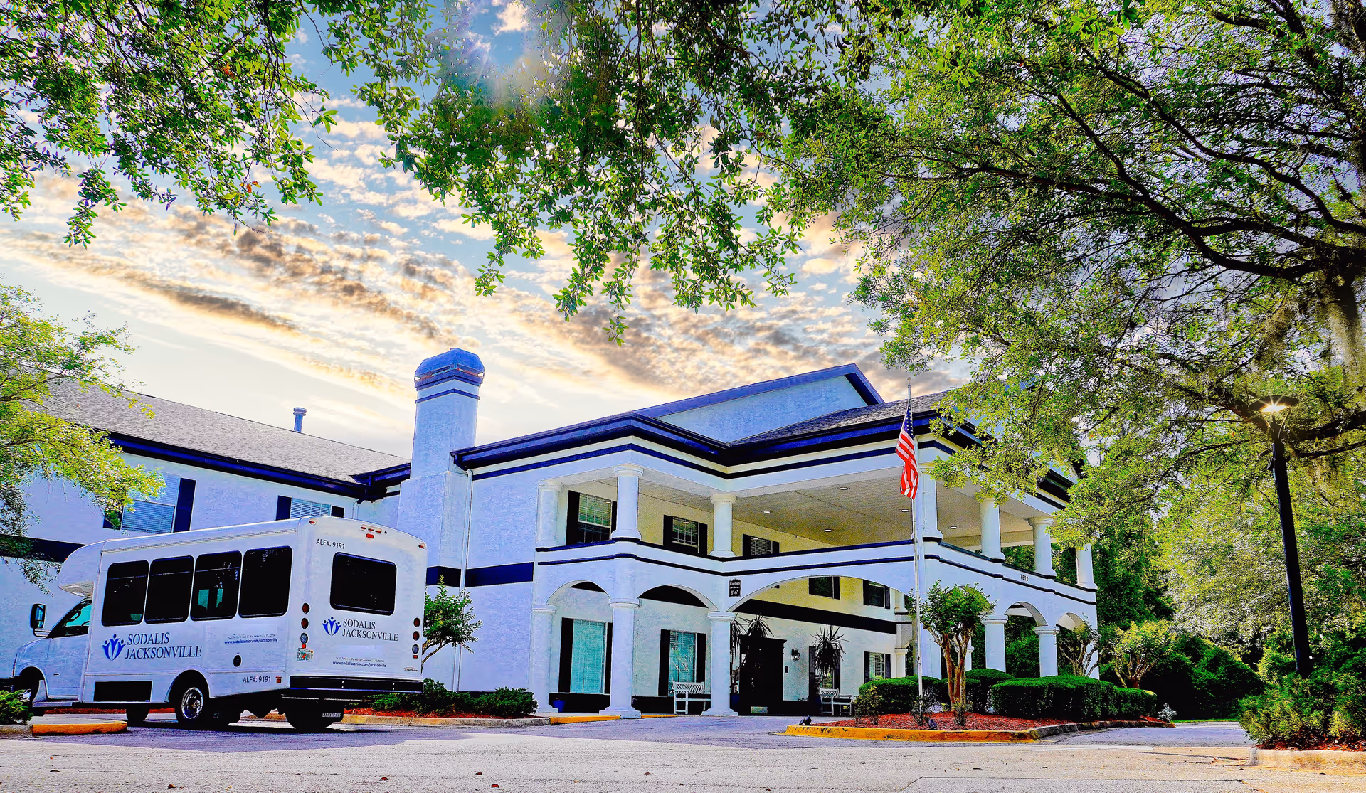 Front exterior of a two-story white senior living building with columns, an American flag, and a shuttle van parked in the driveway.