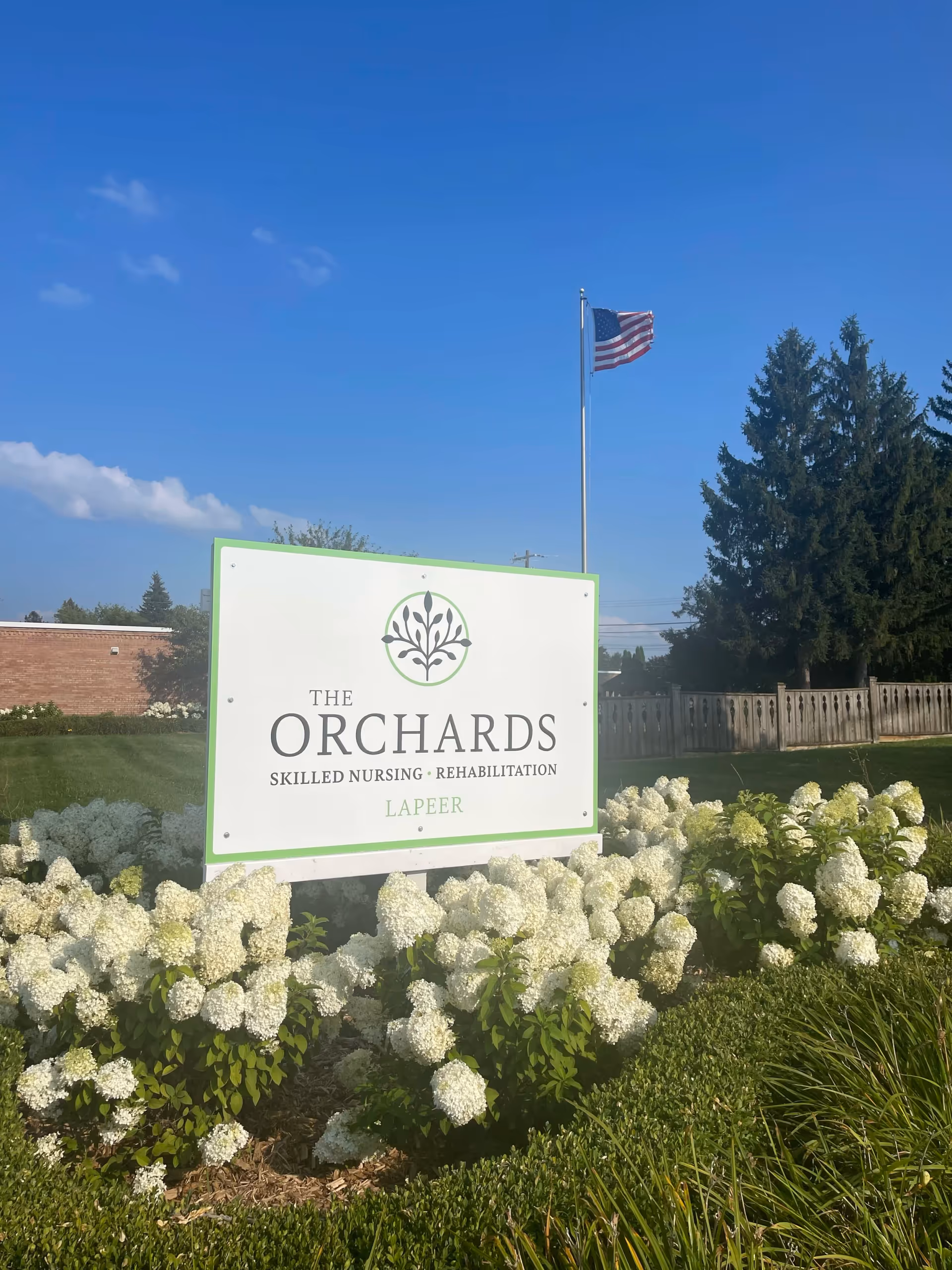 Exterior sign for The Orchards at Lapeer with white flowering plants and an American flag in a clear blue sky.