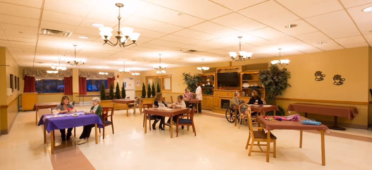 A spacious dining room in a senior living facility with several tables covered with tablecloths. Multiple elderly residents and staff are seated or standing, engaging in conversation. The room is warmly lit with chandeliers, decorated with plants, wall art, and a large wooden entertainment center with a TV and fireplace.