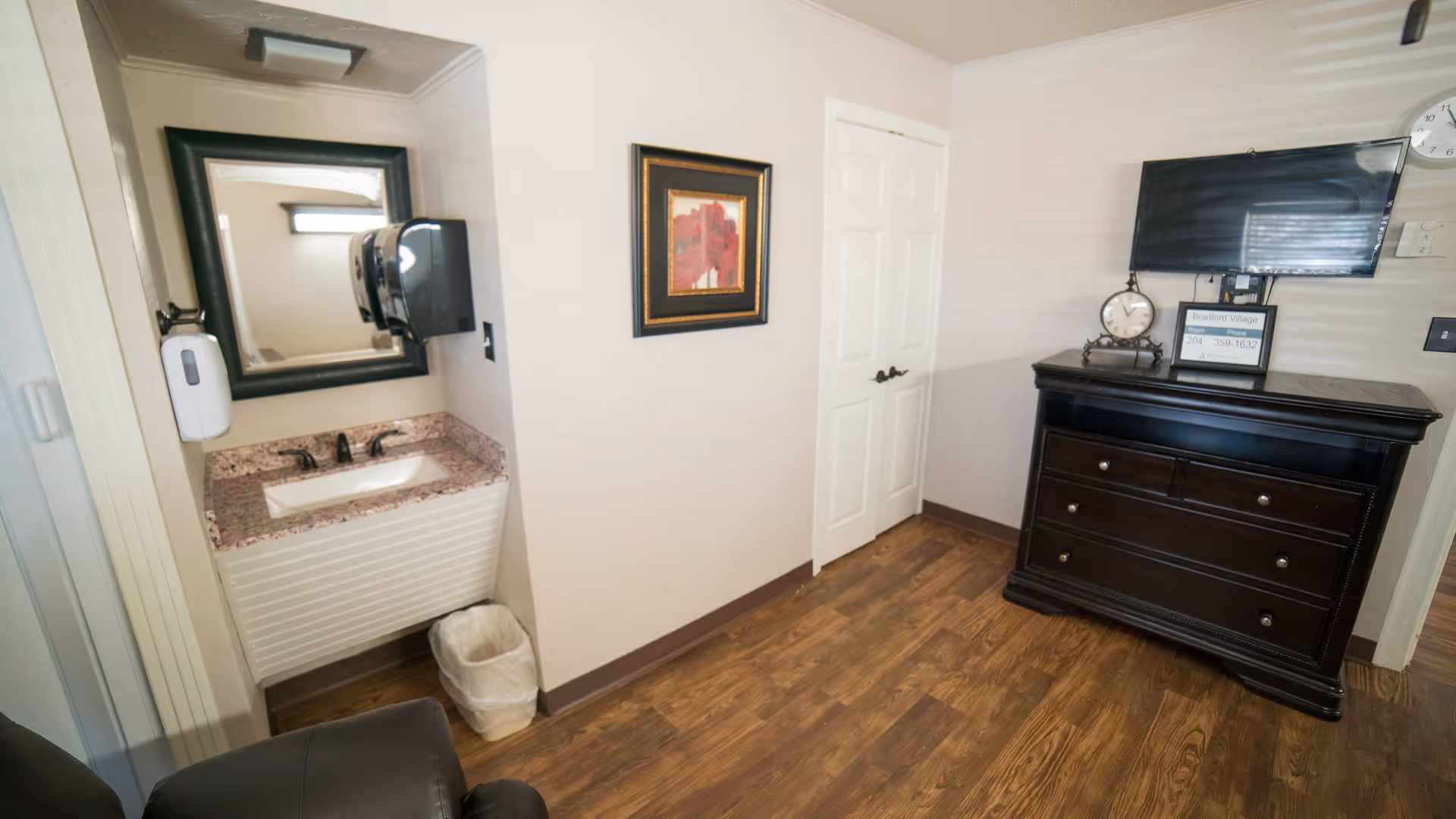 Small senior living room interior showing a vanity sink and mirror, a dresser topped with a TV and clock, and wood floors.