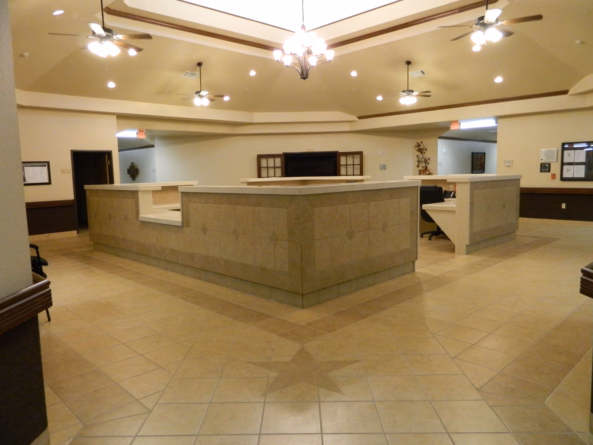 Interior view of a nursing and rehabilitation center reception area with a large tiled counter in the center, beige tiled floor with a star pattern, ceiling fans, and overhead lighting fixtures.