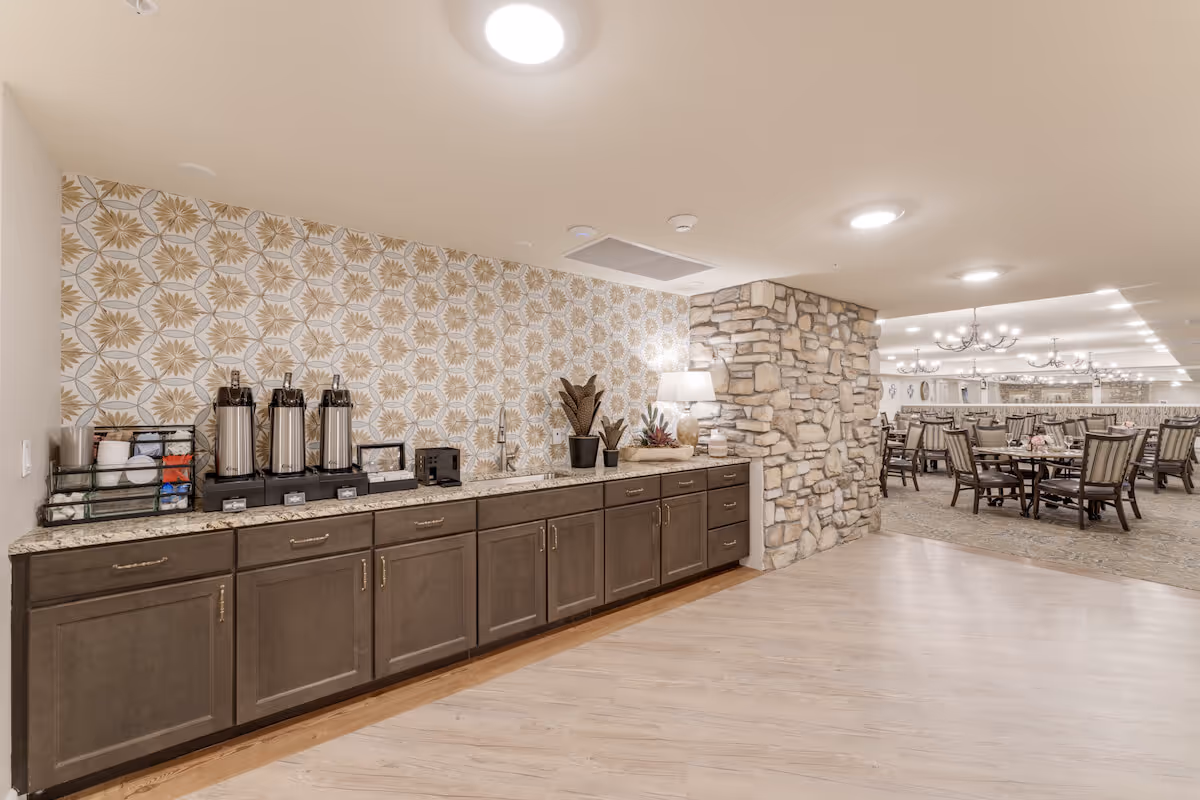 Interior dining area with a long counter and coffee station along a patterned wall, a stone pillar, and multiple dining tables and chairs.