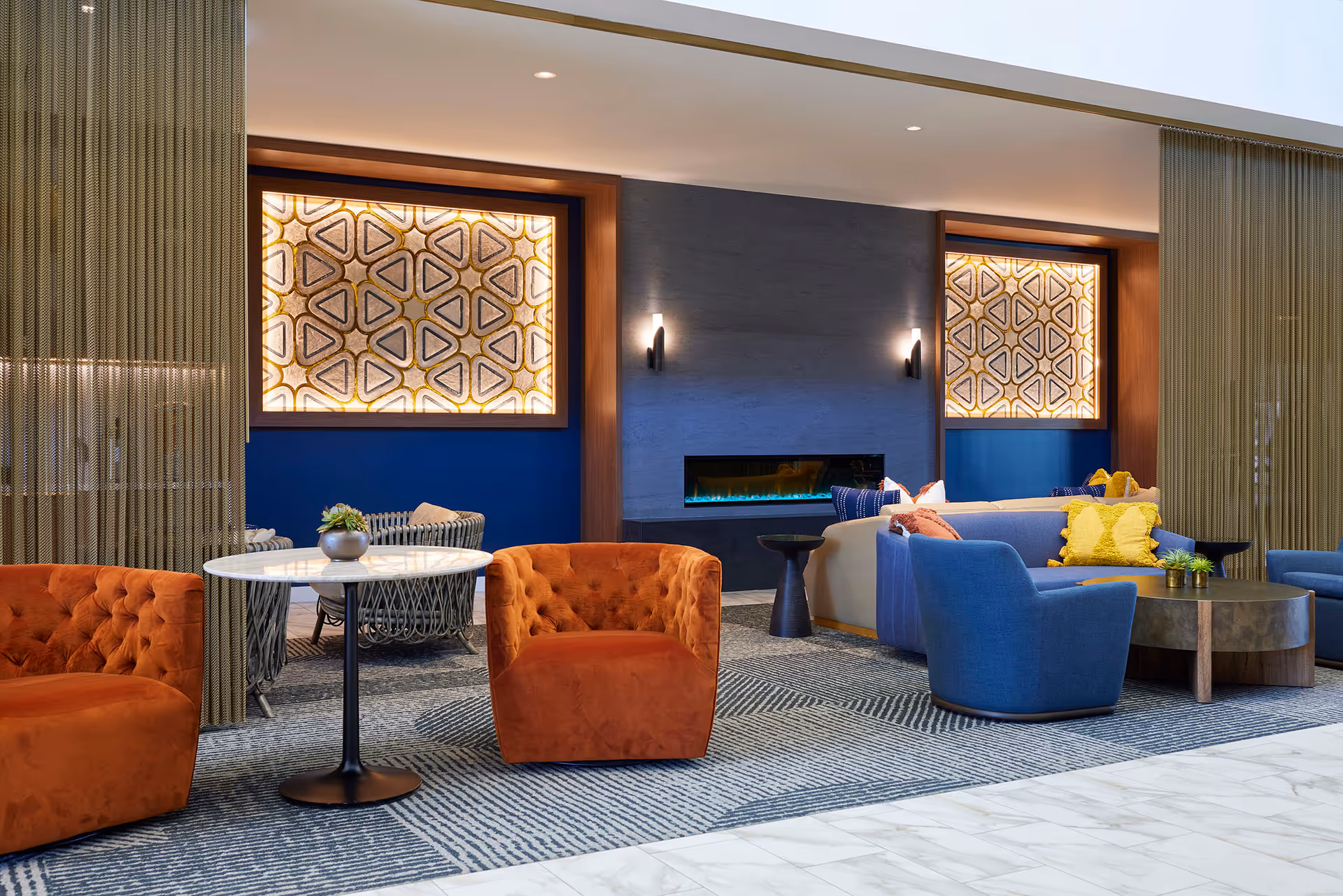 Contemporary communal lounge with orange and blue chairs, sofas, a central coffee table, and a wall-mounted fireplace.